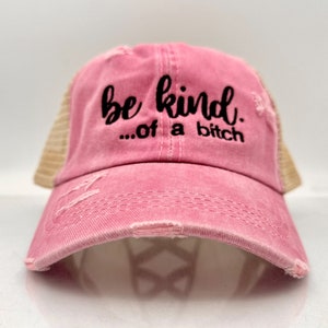May include: A distressed pink trucker hat with a mesh back. The front of the hat features the embroidered phrase "be kind...of a bitch" in black script. The hat has a curved brim and a worn, vintage look.