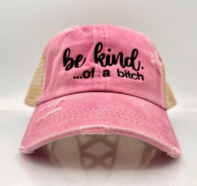 May include: A distressed pink trucker hat with a mesh back. The front of the hat features the embroidered phrase "be kind...of a bitch" in black script. The hat has a curved brim and a worn, vintage look.