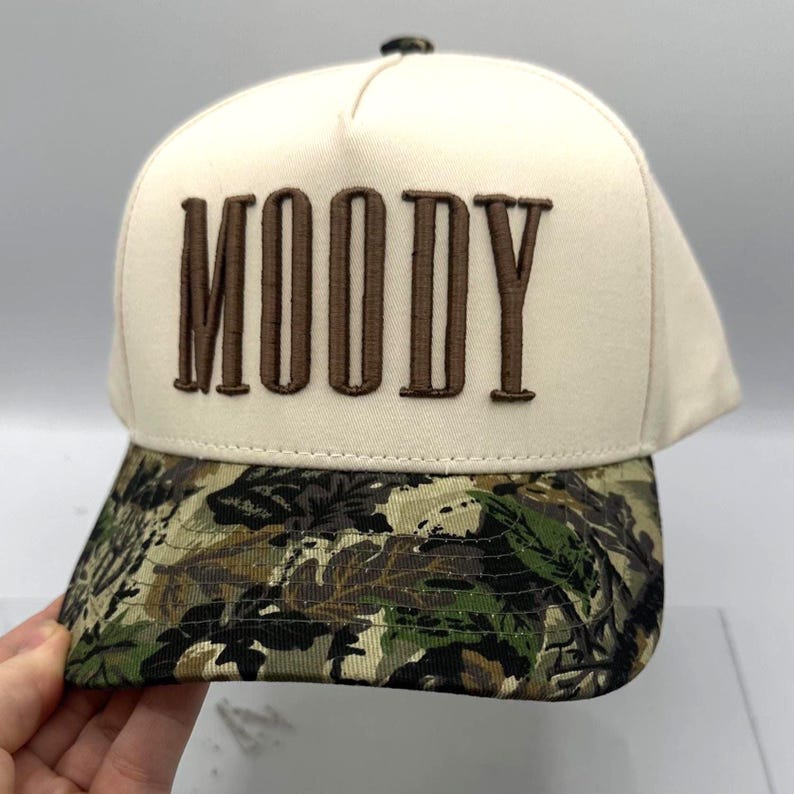 May include: A cream-colored baseball cap with the word "MOODY" embroidered in brown block letters. The brim features a camouflage pattern in shades of green, brown, and black. The cap has a button on top.