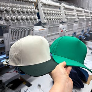 May include: Two five-panel baseball caps, one in off-white with a dark green brim and perforated side panels, and one in solid bright green. Both caps feature a structured crown and a flat brim, presented together in a manufacturing setting.