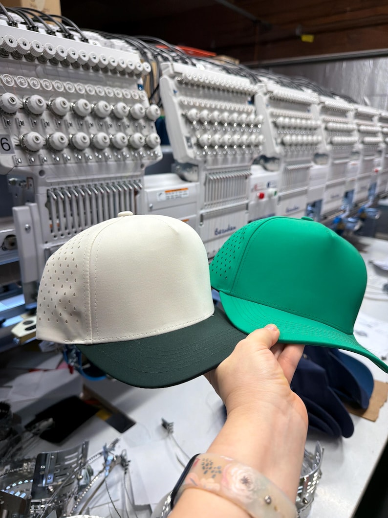 May include: Two five-panel baseball caps, one in off-white with a dark green brim and perforated side panels, and one in solid bright green. Both caps feature a structured crown and a flat brim, presented together in a manufacturing setting.