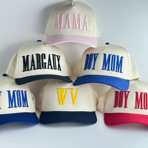 May include: Cream-colored baseball caps with embroidered text in various colors. The caps feature phrases like "MAMA", "MARGAUX", and "BOY MOM", along with a "WV" design. Each cap has a colored brim, including pink, navy, blue, and red.