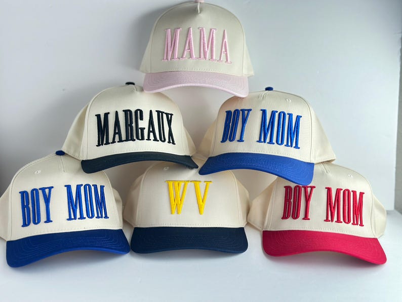 May include: Cream-colored baseball caps with embroidered text in various colors. The caps feature phrases like "MAMA", "MARGAUX", and "BOY MOM", along with a "WV" design. Each cap has a colored brim, including pink, navy, blue, and red.