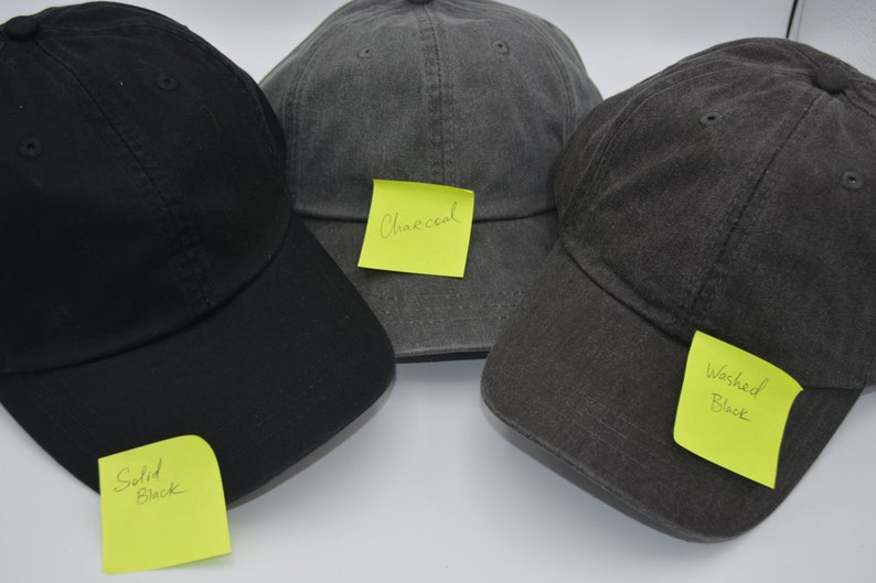May include: Three baseball caps in different shades of grey. The cap on the left is solid black with a note that says "Solid Black". The middle cap is charcoal grey with a note that says "Charcoal". The cap on the right is a washed black with a note that says "Washed Black".