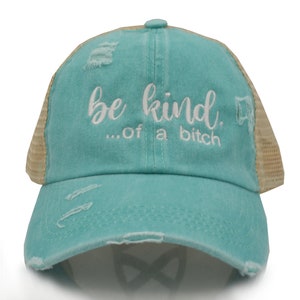 May include: A distressed turquoise baseball cap with a mesh back. The cap features the embroidered phrase "be kind...of a bitch" in white script. The hat has a pre-curved bill and distressed details.