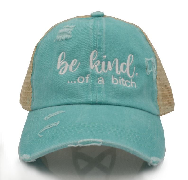 May include: A distressed turquoise baseball cap with a mesh back. The cap features the embroidered phrase "be kind...of a bitch" in white script. The hat has a pre-curved bill and distressed details.