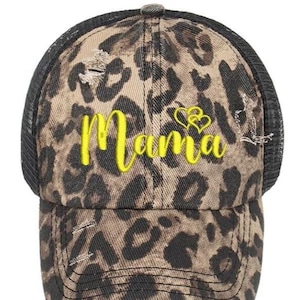May include: A distressed baseball cap with a leopard print pattern in shades of brown and black. The word "Mama" is embroidered in bright yellow across the front, with two yellow hearts above the word. The cap has a black mesh back.