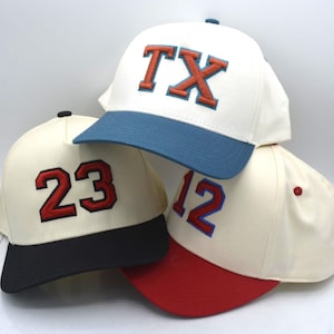 May include: Three baseball caps with cream-colored crowns and contrasting brims. One cap features the letters "TX" in orange and blue, another has the number "23" in red, and the third displays "12" in red. The caps are stacked.