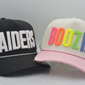 May include: A black baseball cap with white embroidered text reading RAIDERS and a white rope detail, alongside a cream-coloured baseball cap with a light pink brim and multicoloured embroidered text reading BOOZIE in neon shades.