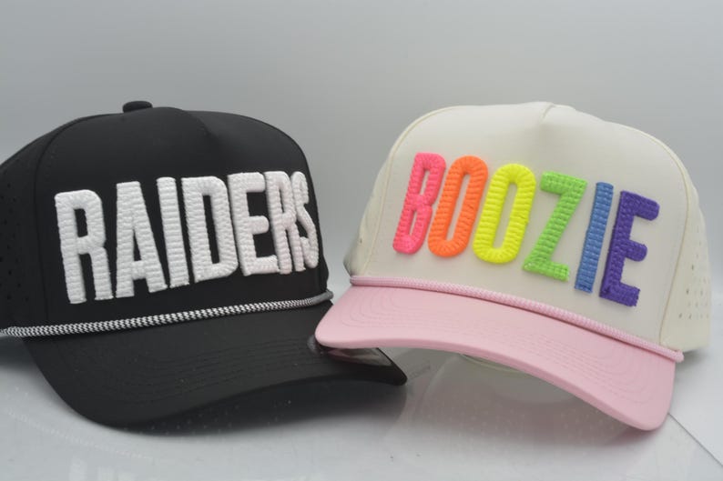May include: A black baseball cap with white embroidered text reading RAIDERS and a white rope detail, alongside a cream-coloured baseball cap with a light pink brim and multicoloured embroidered text reading BOOZIE in neon shades.
