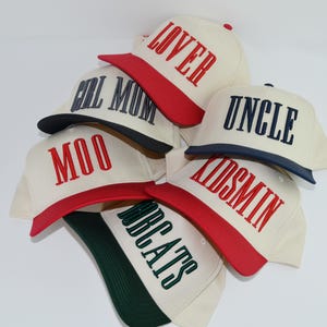 May include: A collection of cream-colored baseball caps with various embroidered words in bold lettering. The caps have colored brims and the words include "LOVER", "FULL MUM", "MOO", "UNCLE", "KIDSMIN", and "BEATS".