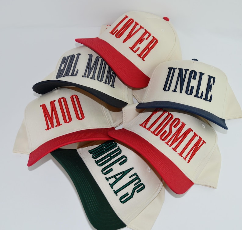 May include: A collection of cream-colored baseball caps with various embroidered words in bold lettering. The caps have colored brims and the words include "LOVER", "FULL MUM", "MOO", "UNCLE", "KIDSMIN", and "BEATS".