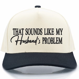 May include: A baseball cap with a cream-colored crown and a black brim. The cap features the text "THAT SOUNDS LIKE MY Husband's PROBLEM" in black lettering, making it a humorous accessory.