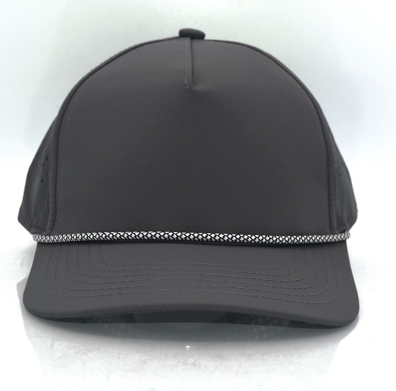 May include: A solid dark grey baseball cap featuring a five-panel construction with a structured crown and a curved brim. A decorative, reflective silver and black braided cord trim runs horizontally across the base of the front crown panel.
