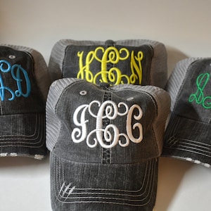 May include: Four gray distressed trucker hats with different colored embroidered monograms. The monograms are KND in blue, HEN in yellow, JCG in white, and SNS in green.