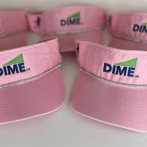 May include: Pink visors with the word "DIME" in navy blue and a green triangle logo. The visors have a silver rhinestone trim and a golf-themed graphic on the side. The visors are made of a soft, breathable fabric.
