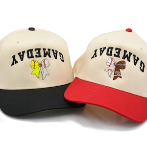 May include: Two beige baseball caps with black lettering that reads "GAMEDAY". One cap has a black brim and the other has a red brim. Both caps feature a colorful embroidered bow with a softball or football in the center.