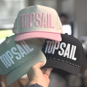 May include: Three trucker hats in the image, each with "TOPSAIL" embroidered on the front. One hat is cream with a pink brim and pink lettering, another is sage green with white lettering, and the third is black with white lettering.