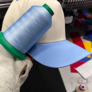 May include: A baseball cap with a light blue brim and a cream-colored crown. A spool of light blue thread rests on the cap, with a green spool base. The cap is likely for embroidery.