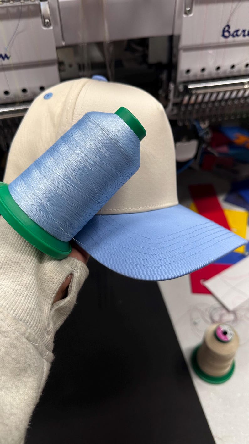 May include: A baseball cap with a light blue brim and a cream-colored crown. A spool of light blue thread rests on the cap, with a green spool base. The cap is likely for embroidery.