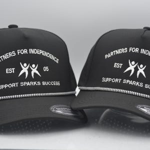 Peut inclure: Deux casquettes de baseball noires avec du texte blanc indiquant "PARTNERS FOR INDEPENDENCE EST 05 SUPPORT SPARKS SUCCESS". Chaque casquette a un d&eacute;tail de corde blanche et argent&eacute;e le long du bord.