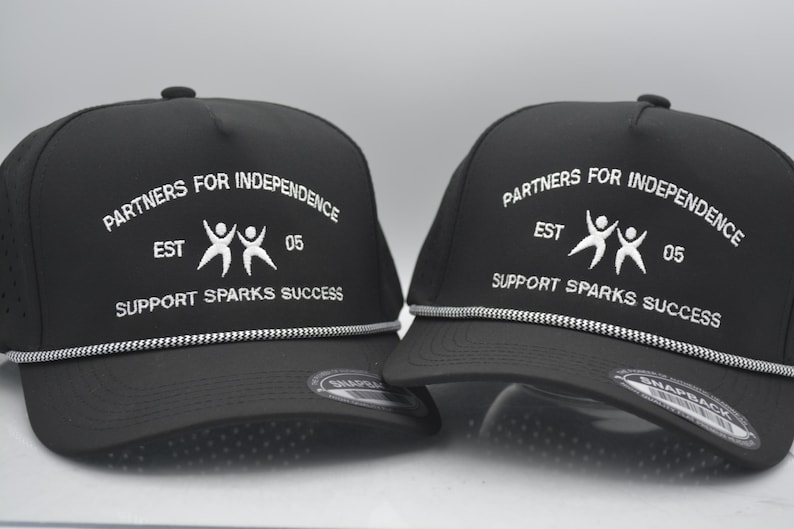 Peut inclure: Deux casquettes de baseball noires avec du texte blanc indiquant "PARTNERS FOR INDEPENDENCE EST 05 SUPPORT SPARKS SUCCESS". Chaque casquette a un d&eacute;tail de corde blanche et argent&eacute;e le long du bord.