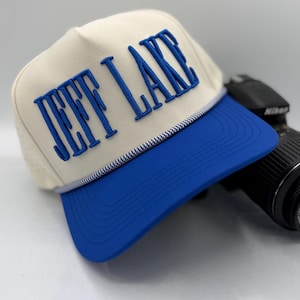 May include: A cream-colored trucker hat with the text "JEFF LAKE" embroidered in blue. The hat features a blue brim and a white and blue rope detail. A black camera is in the background.
