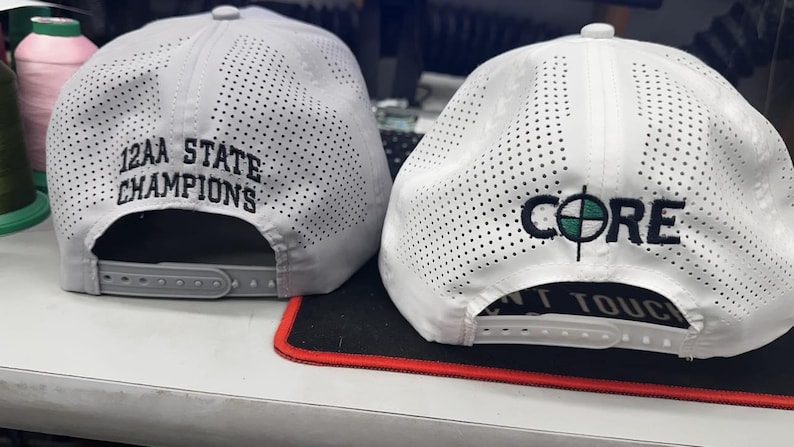 Peut inclure: Deux casquettes de baseball blanches et grises avec des motifs perfor&eacute;s. L'une porte la broderie noire "12AA STATE CHAMPIONS". L'autre affiche "CORE" avec un graphique de cible et le texte "DON'T TOUCH".
