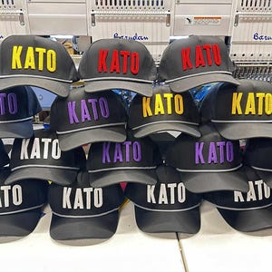 May include: A collection of black baseball caps, each featuring the word KATO embroidered in raised lettering on the front. The caps are available with the text in various colours, including yellow, red, purple, and white, and have a grey brim trim.