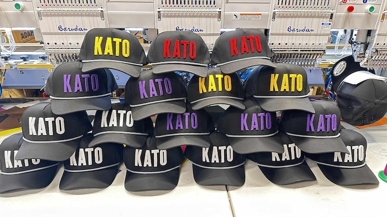 May include: A collection of black baseball caps, each featuring the word KATO embroidered in raised lettering on the front. The caps are available with the text in various colours, including yellow, red, purple, and white, and have a grey brim trim.