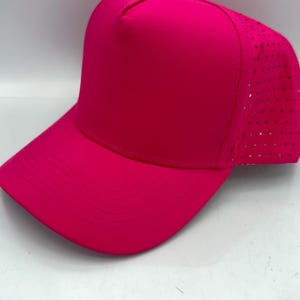 May include: A vibrant hot pink baseball cap featuring a solid front panel and a perforated back panel for ventilation. The cap has a curved brim with stitched detailing and a matching fabric button at the top centre of the crown.