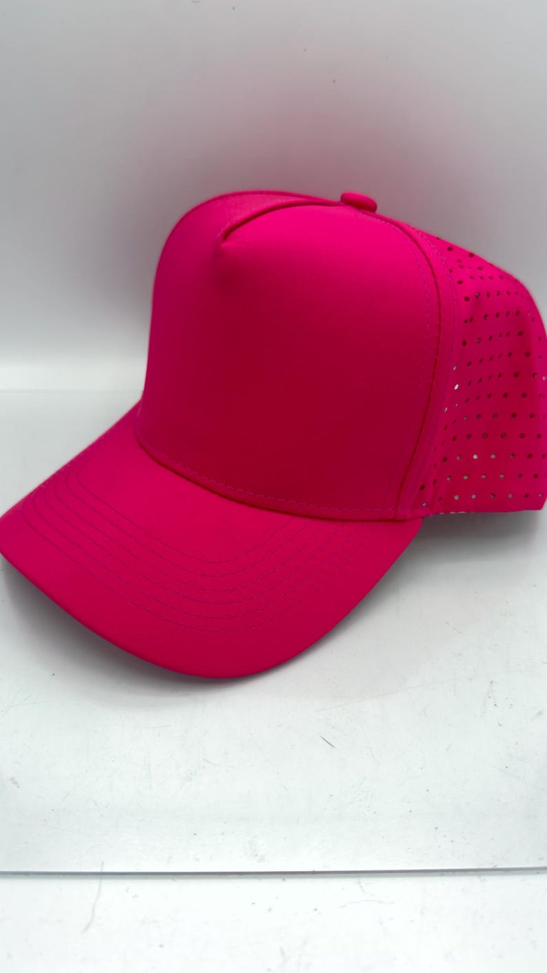 May include: A vibrant hot pink baseball cap featuring a solid front panel and a perforated back panel for ventilation. The cap has a curved brim with stitched detailing and a matching fabric button at the top centre of the crown.
