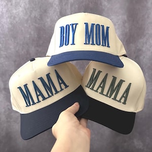 May include: Cream-colored baseball caps with navy blue brims. One cap has "BOY MOM" embroidered in blue, while two others say "MAMA" in blue and green. The caps are stacked, showcasing the design and color scheme.