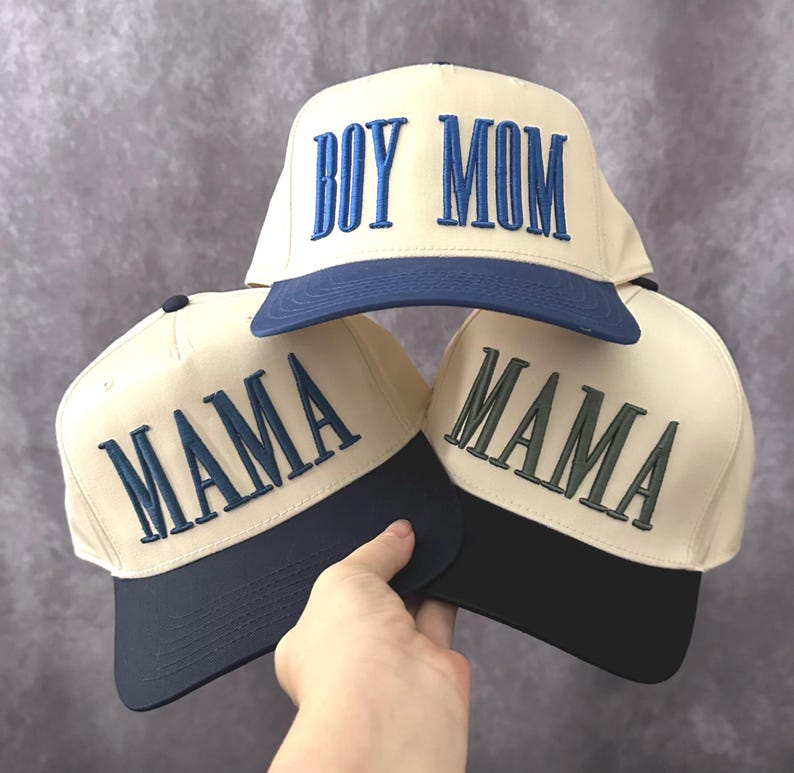 May include: Cream-colored baseball caps with navy blue brims. One cap reads "BOY MOM" in blue embroidery, while two others say "MAMA" in blue and green embroidery. The caps are stacked, with one held by a hand.