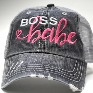 May include: A black and gray distressed trucker hat with a white "BOSS" and pink "babe" embroidered on the front. The hat has a mesh back.