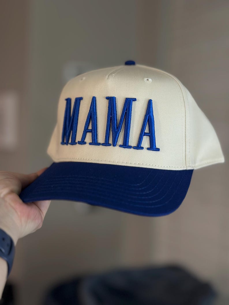 May include: Cream-colored baseball cap with a navy blue brim. The word "MAMA" is embroidered in navy blue block letters across the front of the cap. A classic, stylish accessory.