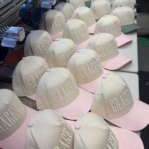 May include: A collection of baseball caps with the word "GRACE" embroidered on the front. The caps have a cream-colored crown and a pink brim. The caps are arranged on a white surface, with an embroidery machine in the background.