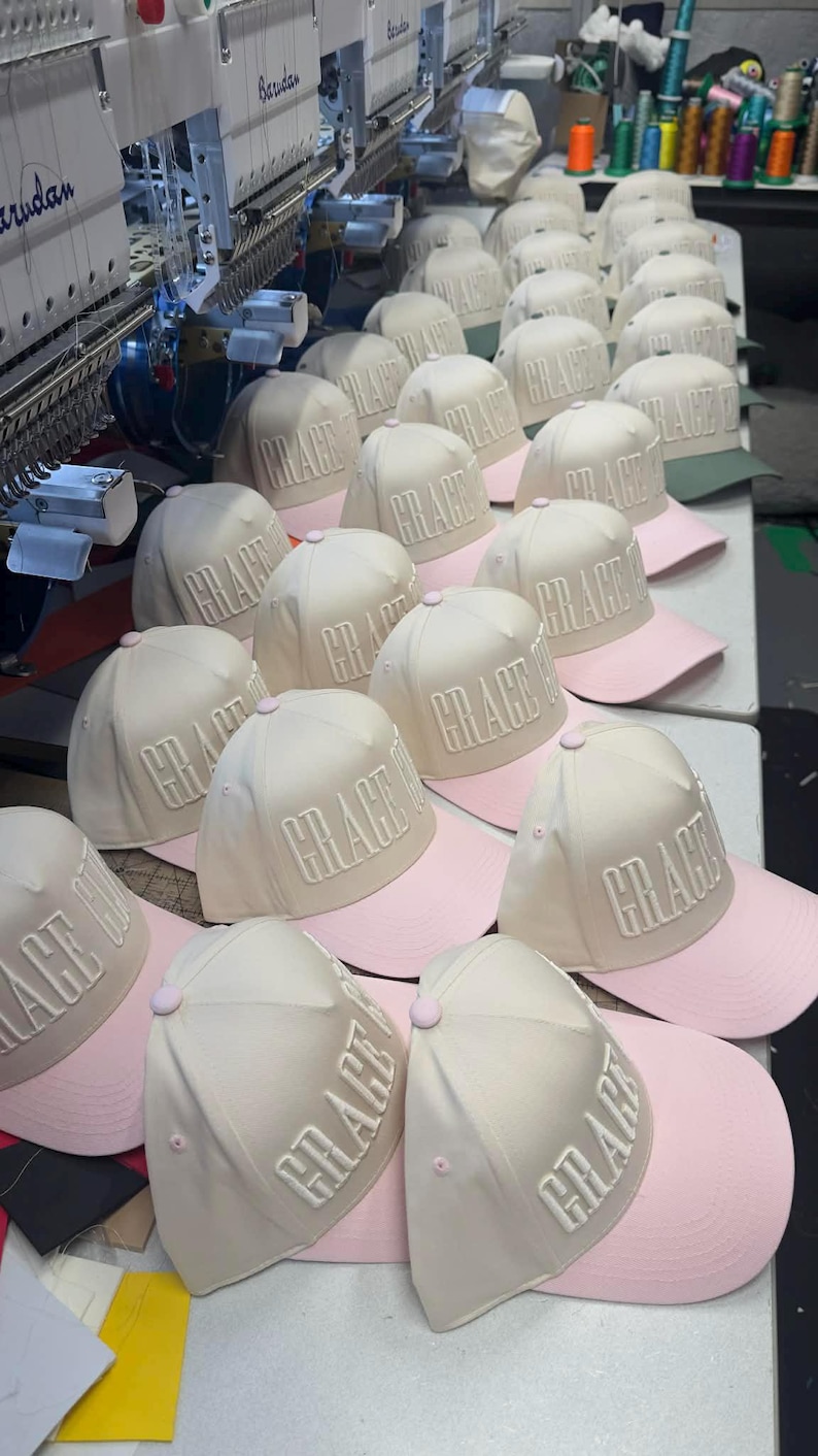 May include: A collection of baseball caps with the word "GRACE" embroidered on the front. The caps have a cream-colored crown and a pink brim. The caps are arranged on a white surface, with an embroidery machine in the background.