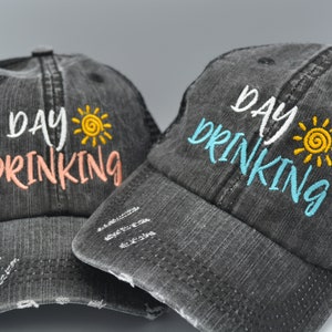 May include: Two black distressed trucker hats with a mesh back. The hats have the text "day drinking" embroidered on the front in white with a yellow sun graphic. One hat has pink embroidery and the other has blue embroidery.