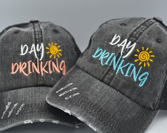 Puede incluir: Dos gorras de camionero negras desgastadas con una malla en la parte posterior. Las gorras tienen el texto "day drinking" bordado en la parte delantera en blanco con un gráfico de sol amarillo. Una gorra tiene bordado rosa y la otra tiene bordado azul.