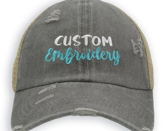May include: A distressed gray trucker hat with a tan mesh back. The hat features the words "CUSTOM Embroidery" embroidered in white and teal. The hat has a pre-worn look with distressed details.