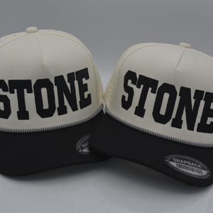 Peut inclure: Deux casquettes snapback cr&egrave;me avec le mot "STONE" en lettres noires en gras. Les chapeaux ont une visi&egrave;re noire et un d&eacute;tail cousu blanc. Le dessus du chapeau comporte de petits trous de ventilation.