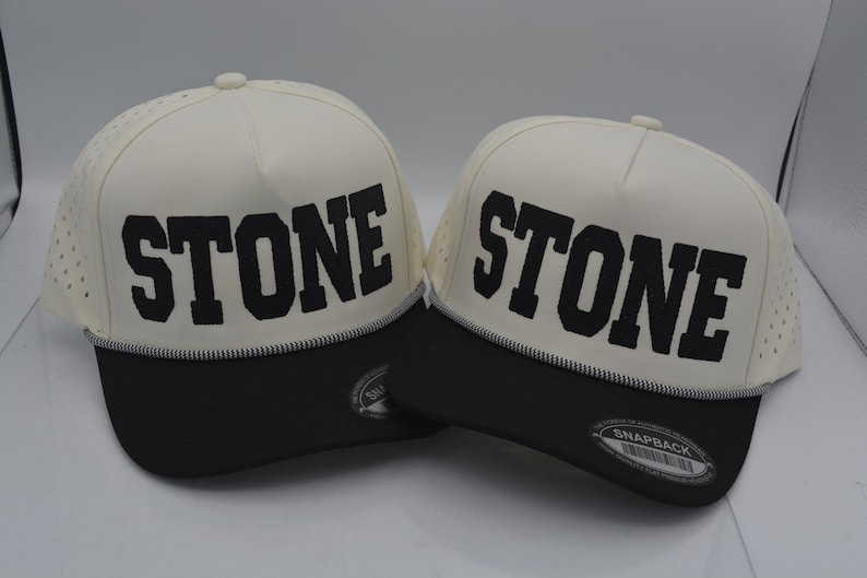 Peut inclure: Deux casquettes snapback cr&egrave;me avec le mot "STONE" en lettres noires en gras. Les chapeaux ont une visi&egrave;re noire et un d&eacute;tail cousu blanc. Le dessus du chapeau comporte de petits trous de ventilation.