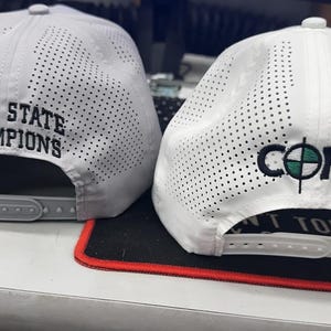 May include: Two white baseball caps with black embroidered text. The cap on the left says "12AA STATE CHAMPIONS". The cap on the right says "CORE" with a green and white logo.