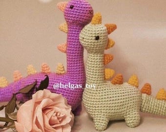 May include: Two crocheted dinosaur toys, one purple and one green, with orange accents. The dinosaurs are sitting on a pink rose.