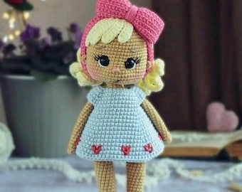 Könnte beinhalten: Eine gehäkelte Puppe mit blonden Haaren, einer rosa Schleife und einem hellblauen Kleid mit roten Herzen. Die Puppe steht auf einer Holzoberfläche. "Tina" @kukla_kropa_toys