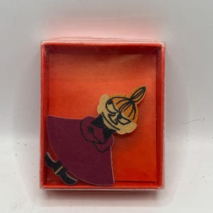 May include: A wooden figurine of a character wearing a purple robe and a yellow hat with a black face. The figurine is in a red box with a clear plastic cover.