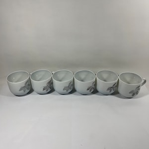 May include: Six white ceramic mugs with a gray leaf design. The mugs are arranged in a row on a white surface.