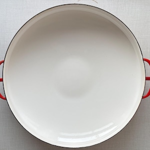 May include: A large, round, white enamelled cast iron pan with a black rim and bright red handles. The interior is a glossy white, and the handles are curved for easy gripping. This cookware is ideal for cooking.
