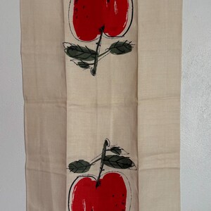 May include: A rectangular beige tea towel featuring two vertically aligned red apple illustrations with black outlines and dark green leaves. The apples are rendered in a painterly style, and the text "USVA" is printed in small black letters at the bottom.